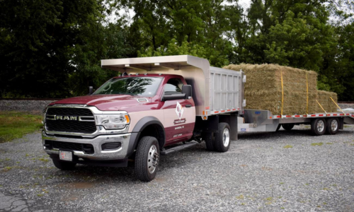 Red Ram truck with a trailer carrying hay bales on a gravel road.